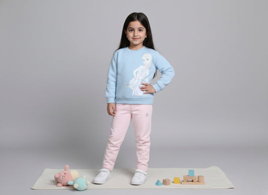 A young girl wearing a baby blue sweatshirt with a frozen design and pink pants, standing on a gray background with toys around.