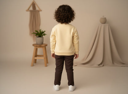 A child wearing a beige sweatshirt and dark pants standing in a neutral-colored room.