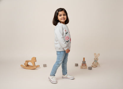 A young girl in a light gray sweatshirt with floral designs, standing in a neutral setting with toys around.