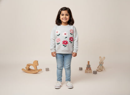 A girl wearing a floral gray sweatshirt and jeans is standing in a neutral setting with toys.