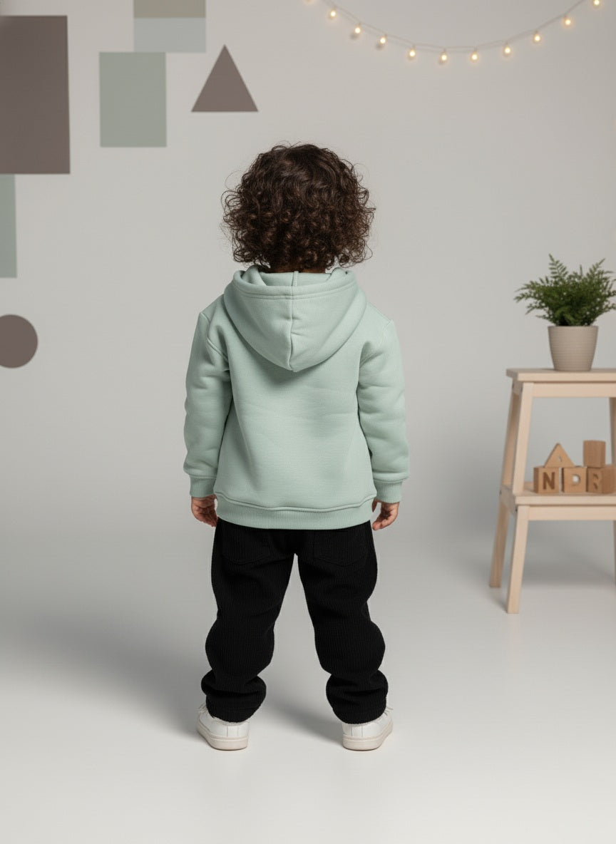 A child wearing a mint green hoodie and black pants is standing in a room with geometric wall designs and a small table.