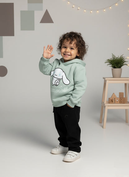 Child wearing a mint green hoodie with a white graphic, standing in a room with a geometric wall design and a small table with plants.