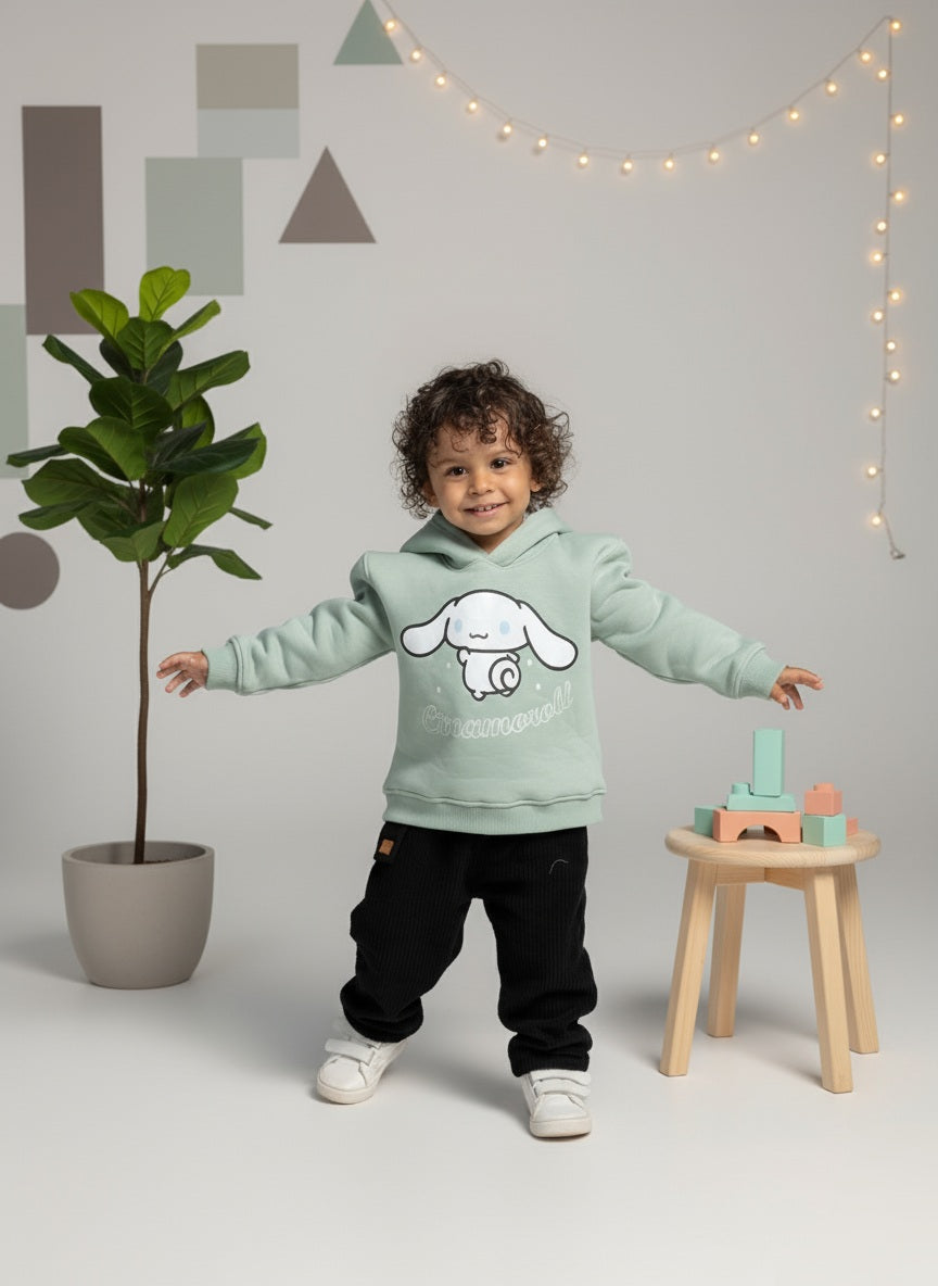 Child wearing a mint green hoodie with a Cinnamoroll design, standing in a room with a plant and a wooden stool.