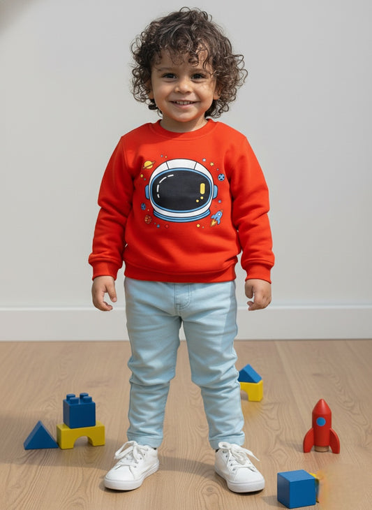 Child wearing an orange sweatshirt with an astronaut design, standing on a wooden floor with toys around.