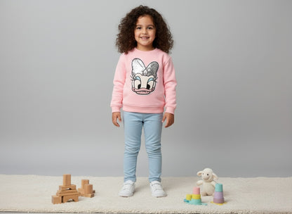 A girl wearing a pink sweatshirt with a daisy patch, standing on a light surface with toys around.